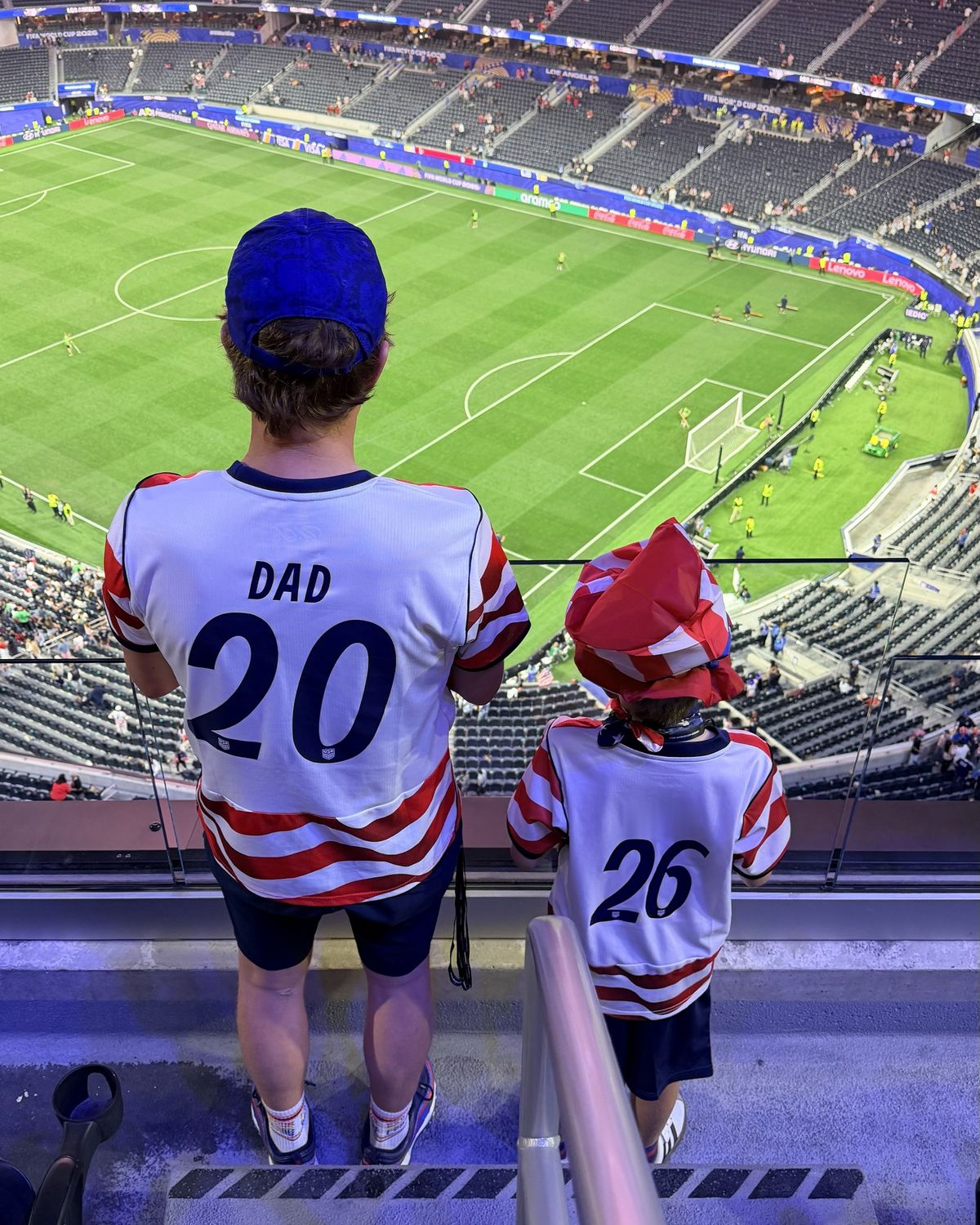 Two fans from behind, a father in a DAD 20 jersey and his son, looking down at the pitch from the top of a World Cup stadium.